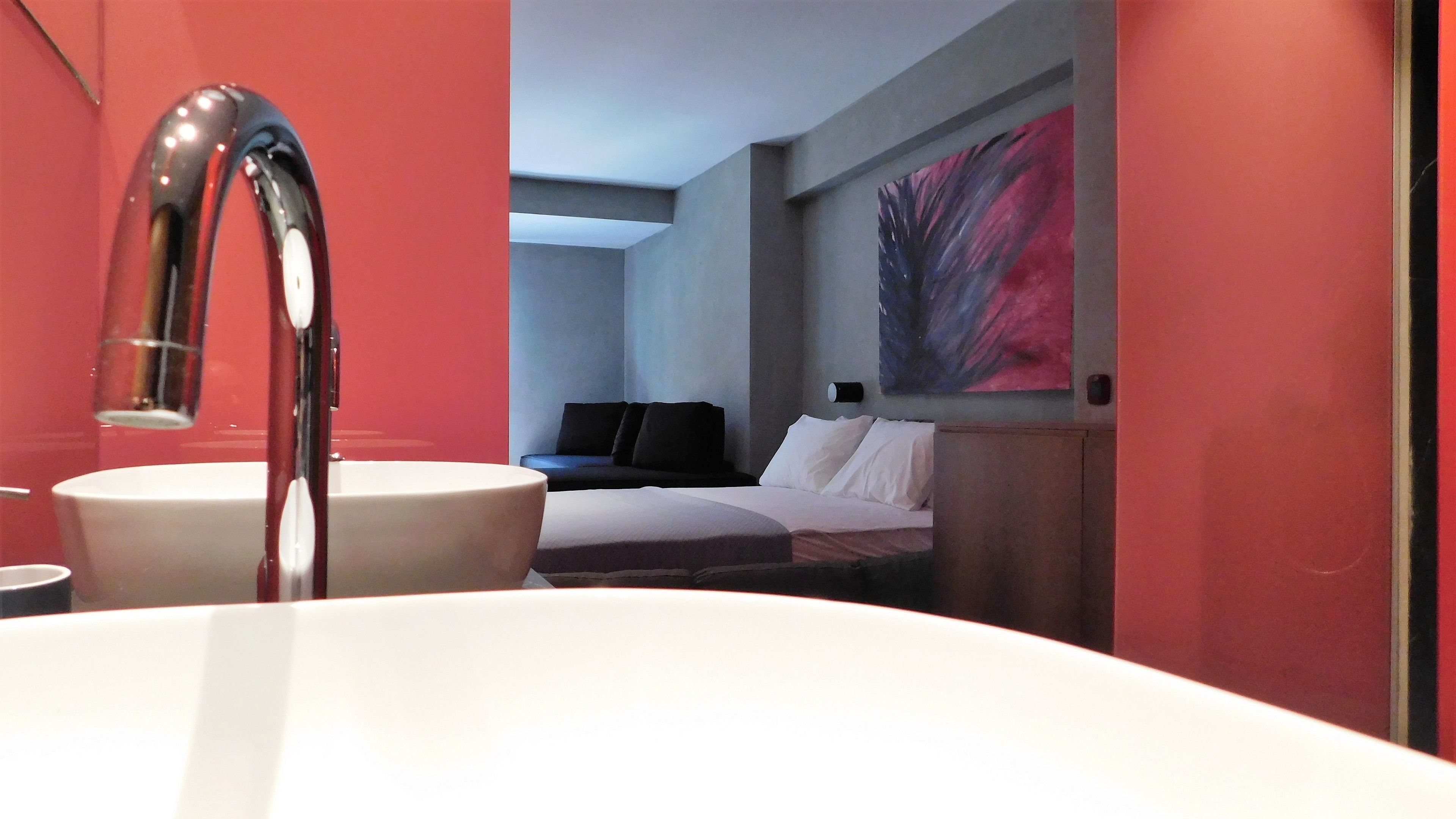 A modern bathroom with a white sink, a mirror, and a bed with white bedding in the background.