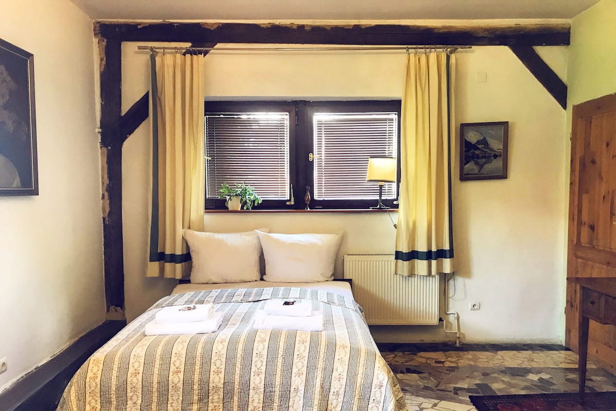 A bedroom with a bed, two windows with blinds, a radiator, a wooden ceiling, and a tiled floor.