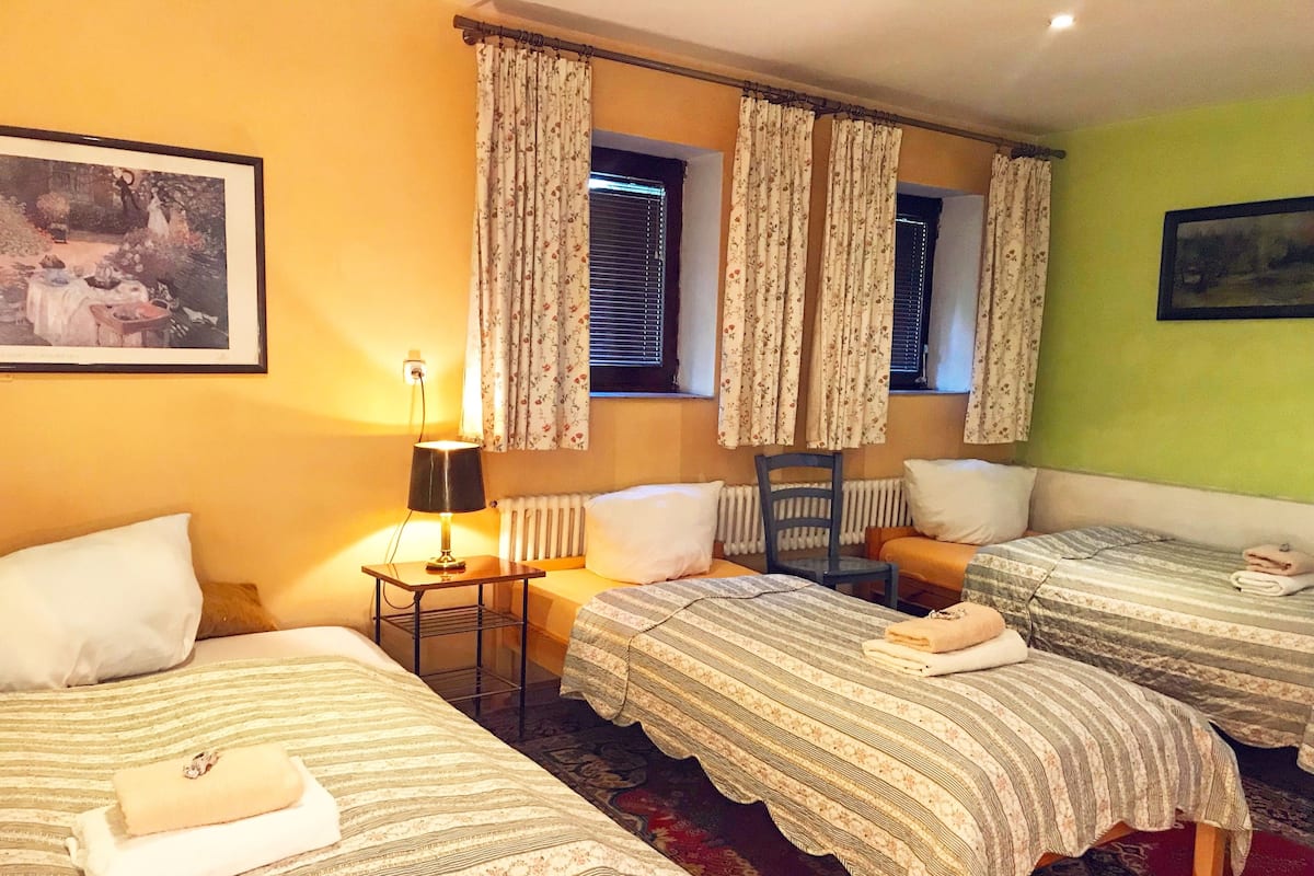 A hotel room with three beds, a small table with a lamp, a chair, and a radiator.