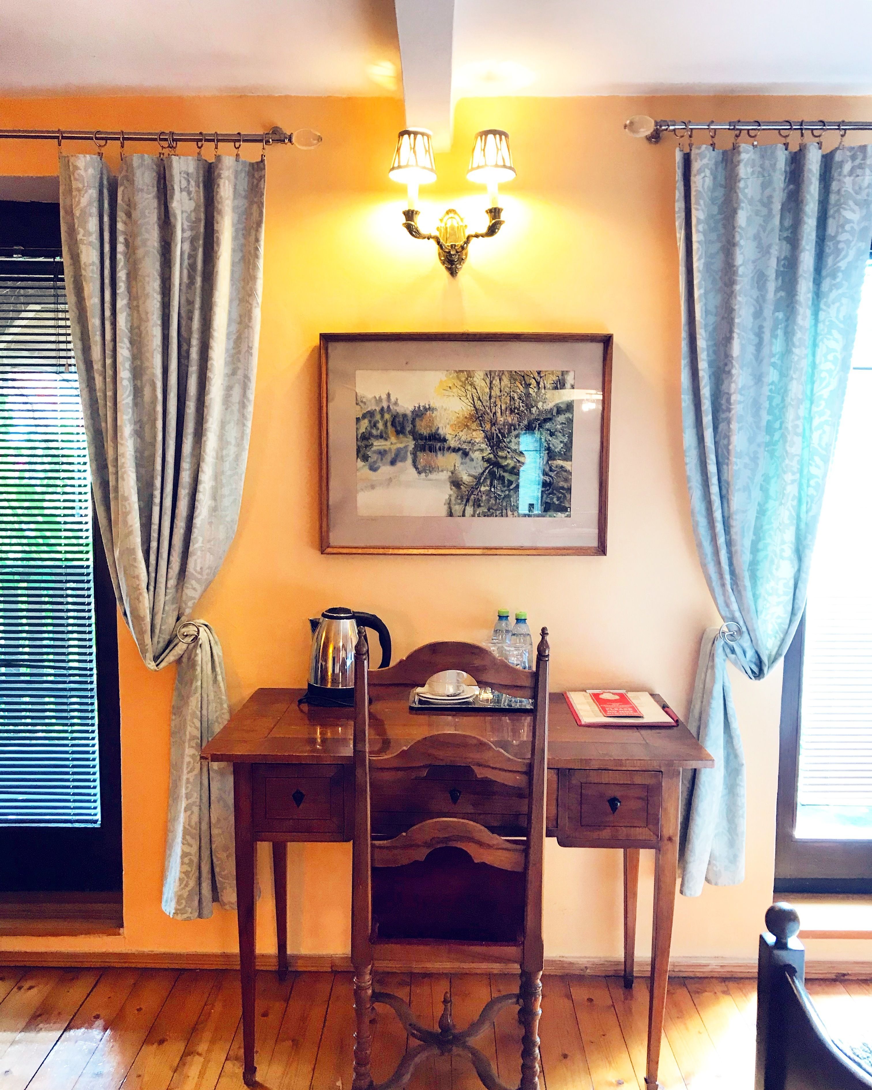 A wooden desk with a kettle, a framed painting, and a chair in a room with wooden flooring and curtains.
