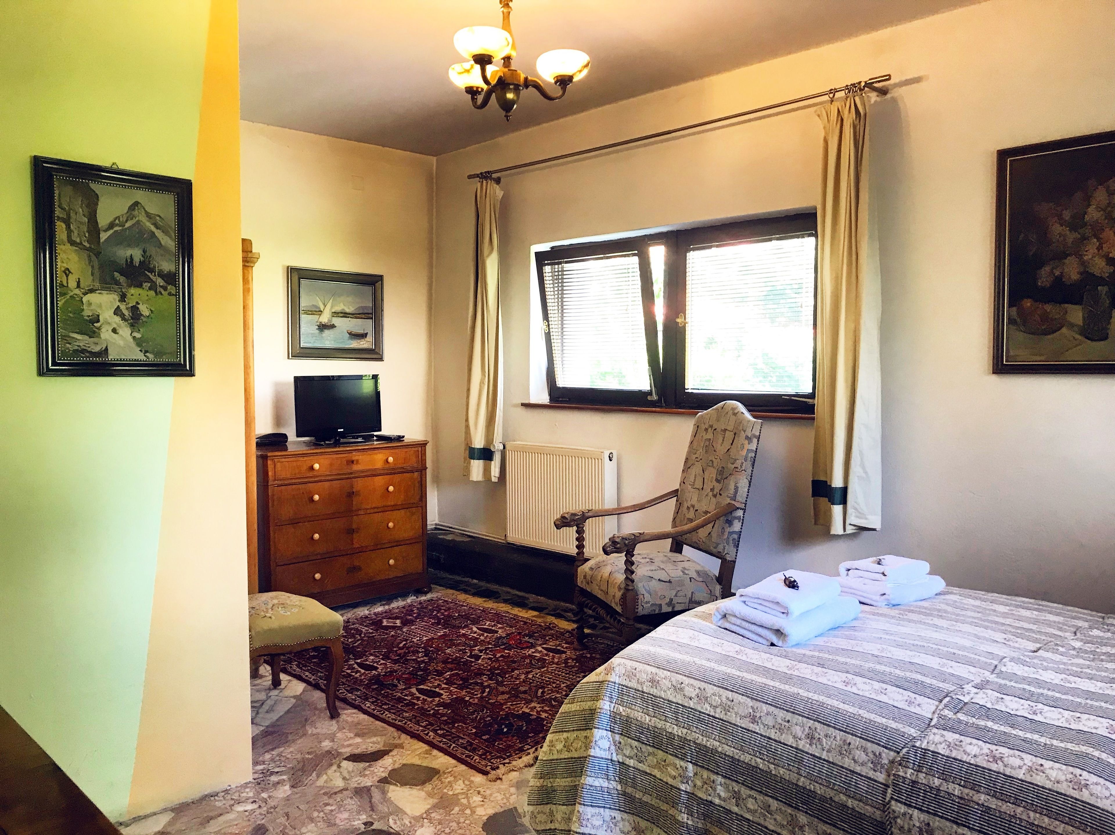 Un dormitorio con cama, una silla, tocador, televisor y ventana con cortinas.
