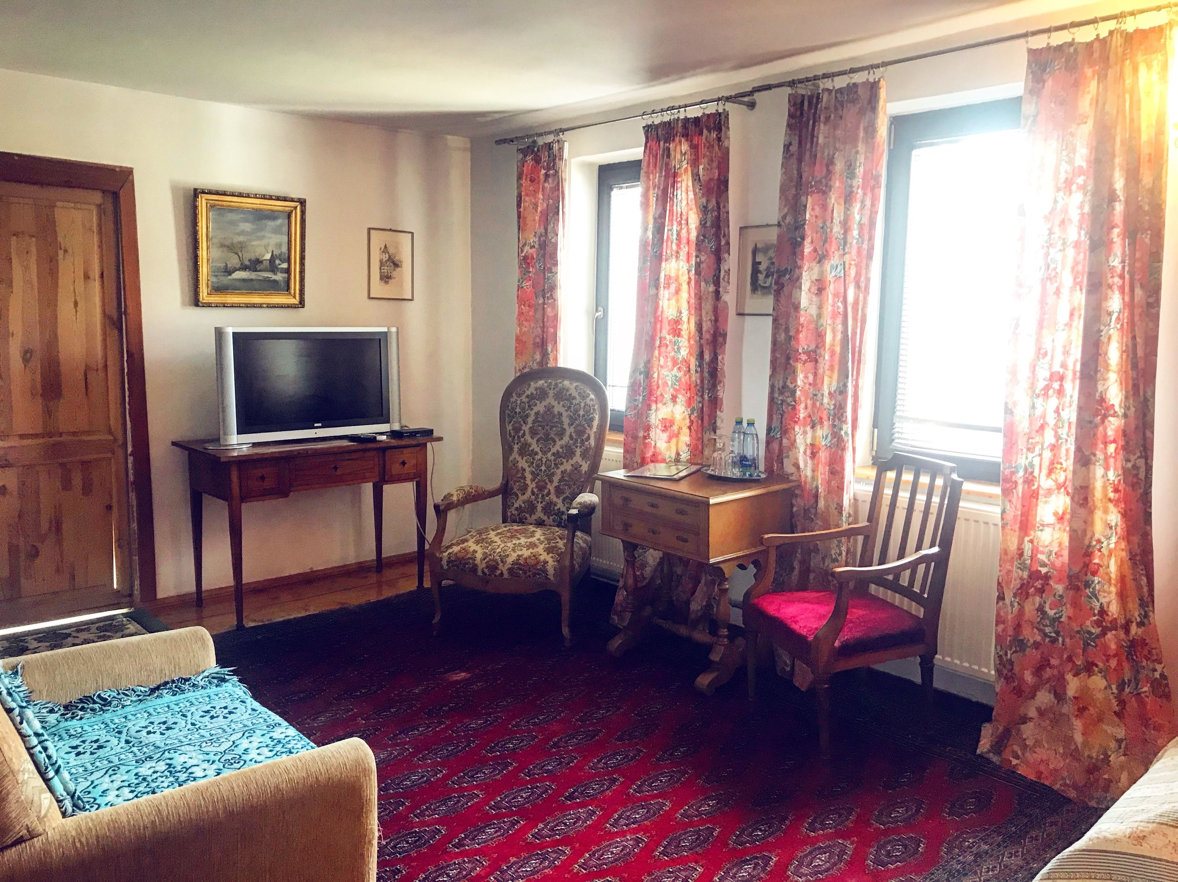 A room with a television on a wooden stand, a patterned armchair, a desk with a chair, and a red patterned carpet.