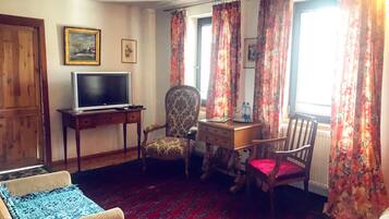 A room with a television on a wooden stand, a patterned armchair, a desk with a chair, and a red patterned carpet.