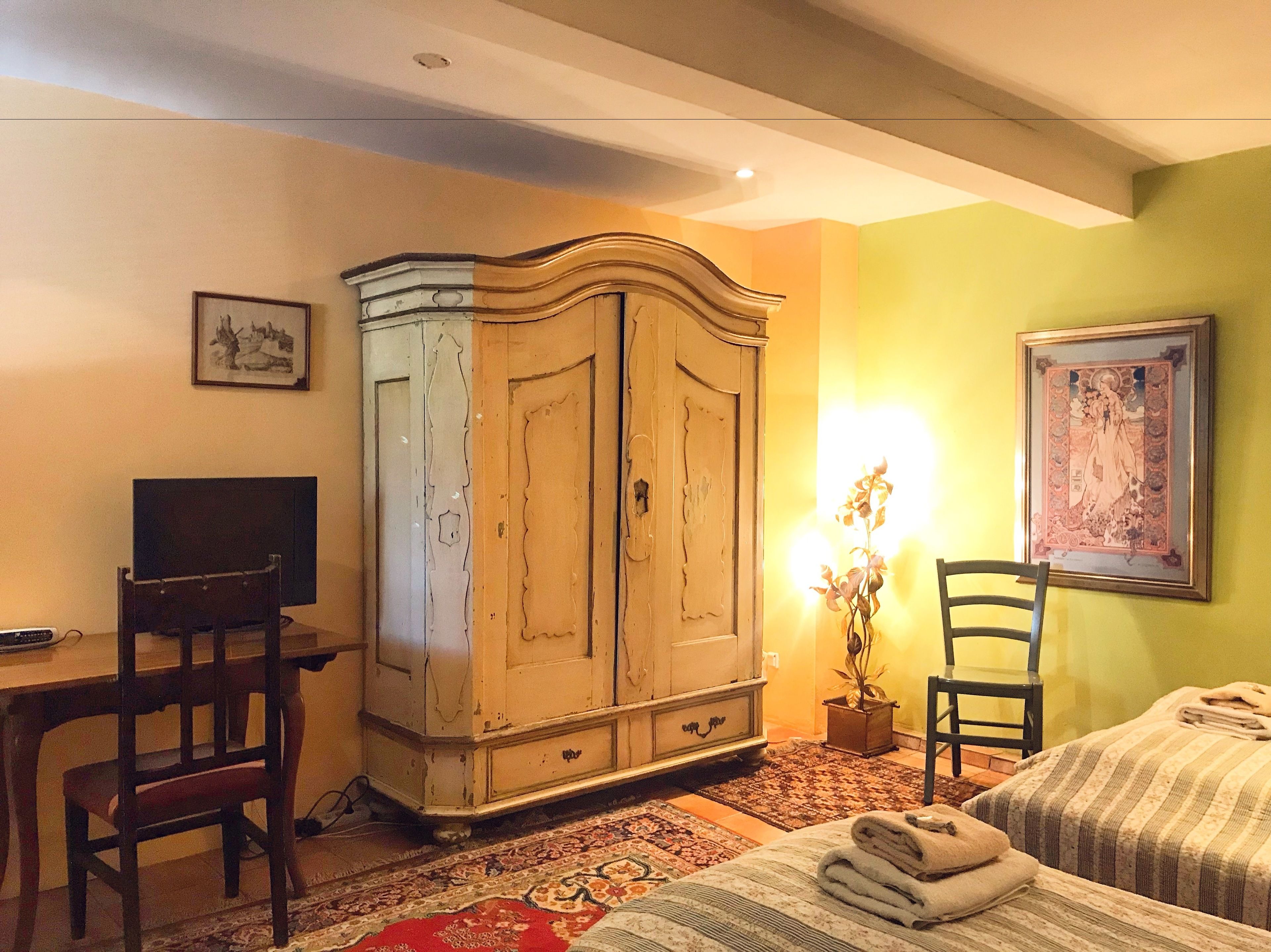 Una habitación con un armario de madera, un escritorio con una computadora, una silla, una cama con una colcha a rayas y un cuadro en la pared.
