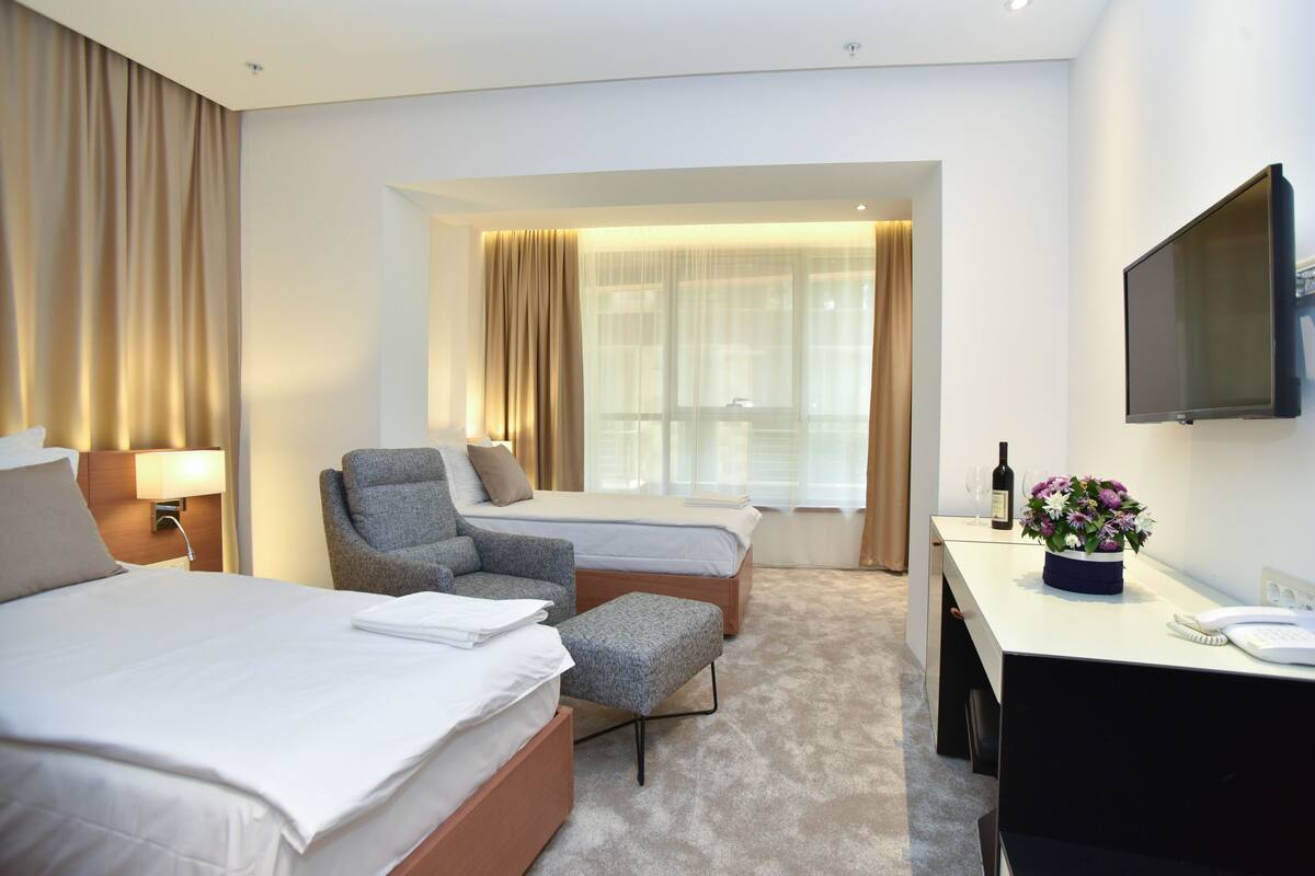 A hotel room with two beds, a desk with a telephone, a TV, and a window with curtains.