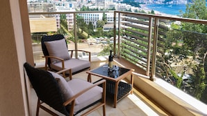 Un balcon avec deux fauteuils en osier, une petite table avec une bouteille, et une vue sur le paysage urbain de la côte.