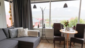 Una sala de estar moderna con un sofá gris, dos sillas, una mesa redonda pequeña y una lámpara de pie. Un ventanal ofrece vistas de un edificio rojo y montañas.