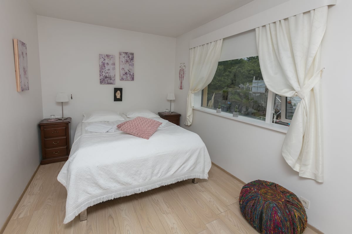 A bedroom with a bed, two bedside tables, a window with curtains, and a small stool.