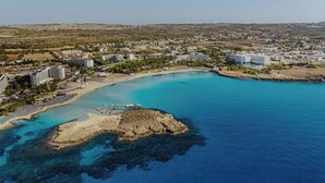 An aerial view of a coastal resort with a sandy beach, clear blue waters, and a rocky outcrop. - Nissiblu Beach Resort (Ayia Napa)