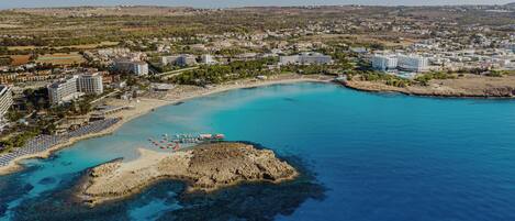 An aerial view of a coastal resort with a sandy beach, clear blue waters, and a rocky outcrop.