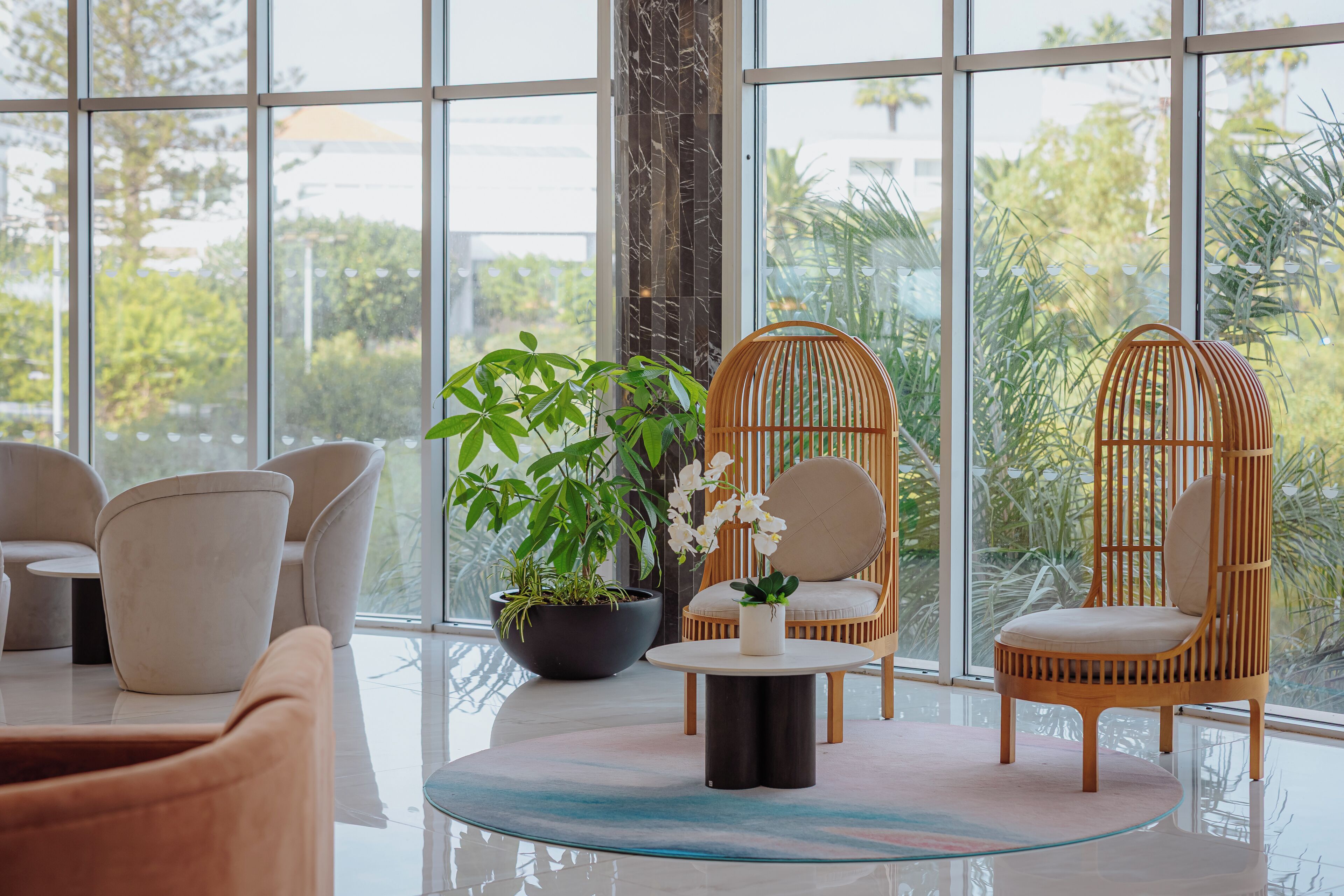 Un interior moderno con ventanas amplias, sillas de mimbre, una mesa redonda de vidrio y una planta en maceta.