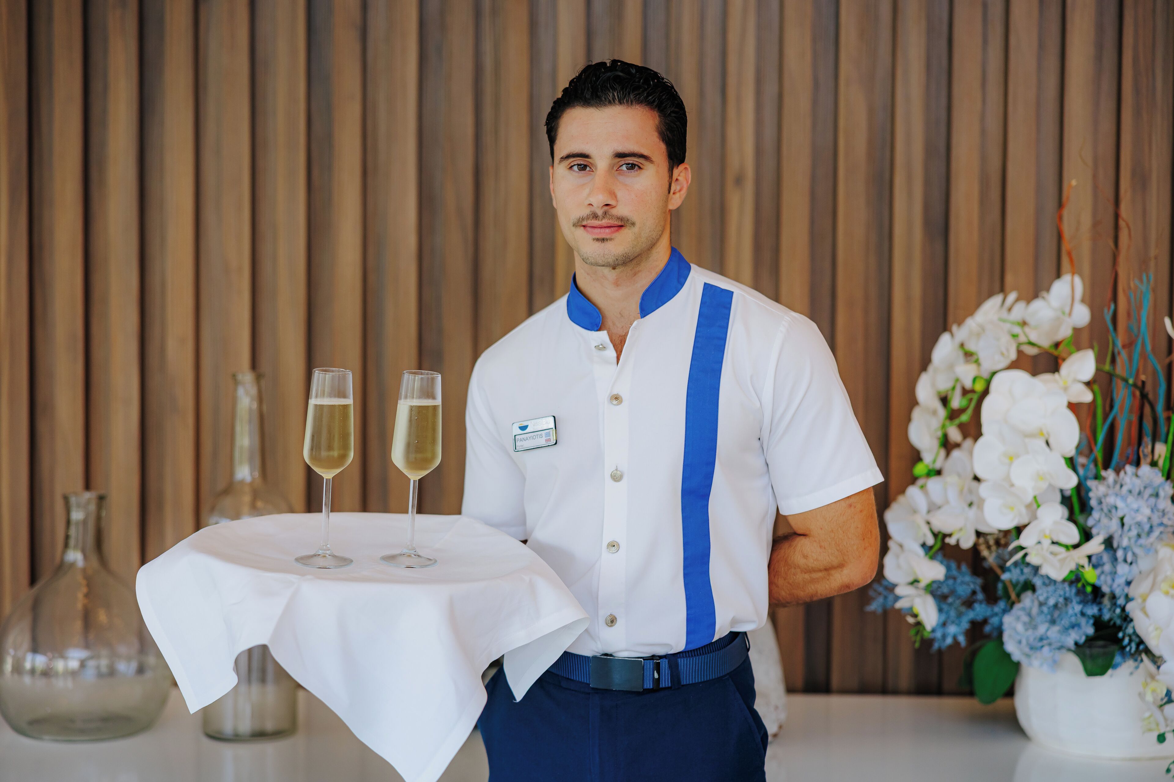 A person in a white shirt with blue stripes holding a table with two glasses of wine.