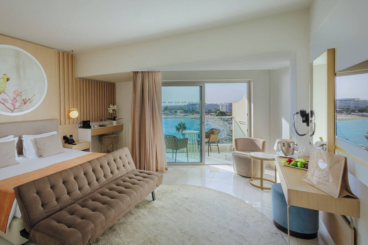 A hotel room with a bed, a sofa, a desk, a phone, a plant, and a view of the beach.