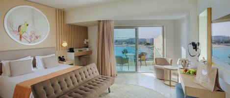 Habitación de hotel con cama, sofá, escritorio, teléfono, planta y vista a la playa.