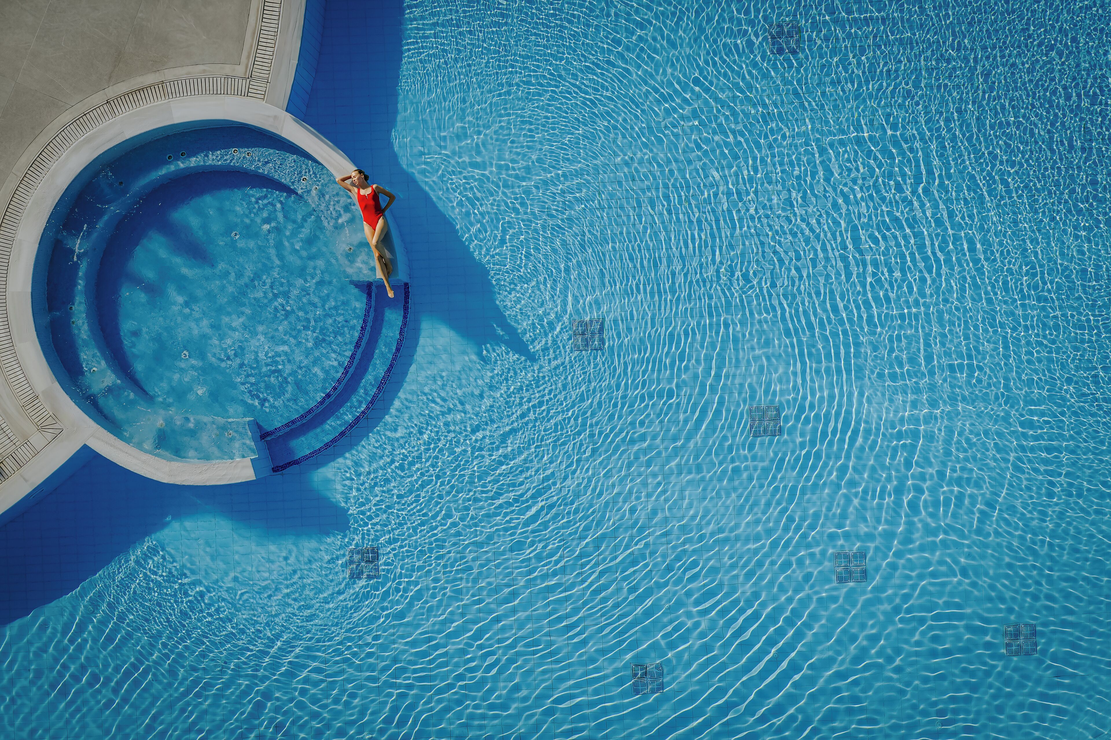An aerial view of a swimming pool with clear water, a hot tub, and a person in a red swimsuit.