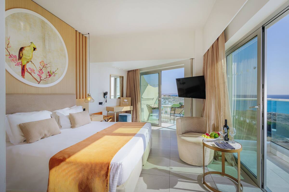 A hotel room with a large bed, a TV, a balcony with a table and chairs, and a view of the sea.