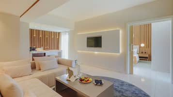 Una sala de estar moderna con sofá, mesa de centro y televisor. Se observa una zona de cocina con paneles de madera y una mesa de comedor.