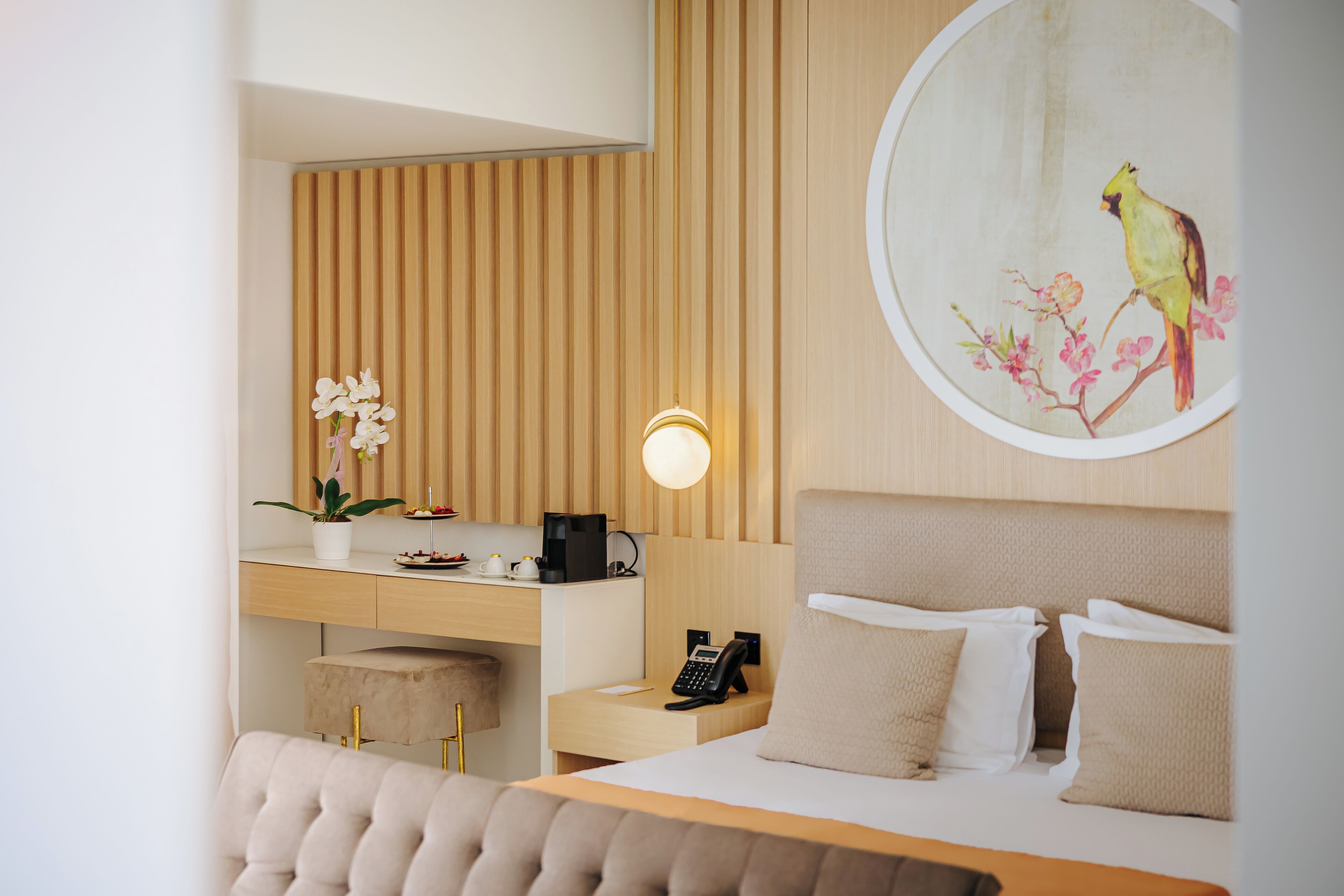 A hotel room with a bed, a desk with a computer, a telephone, a wall-mounted light, and a painting of a bird.