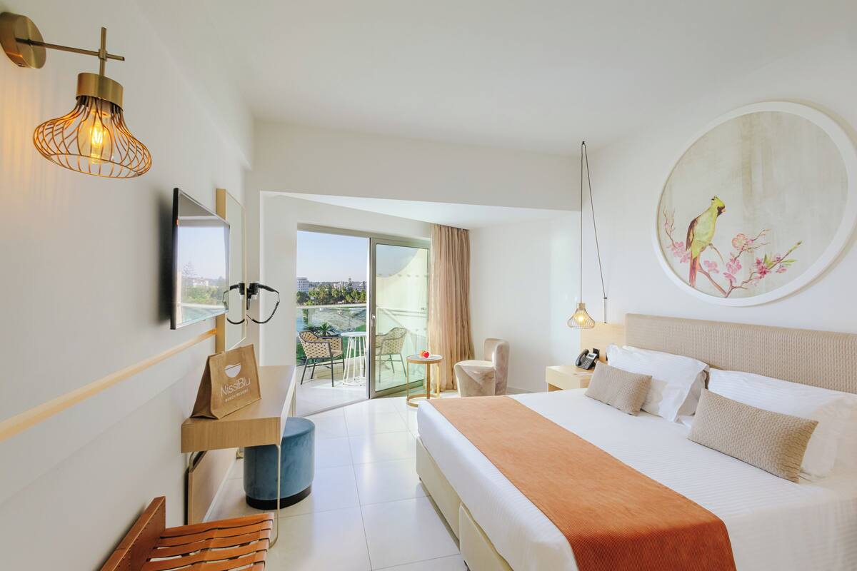 A hotel room with a bed, a desk, a chair, a balcony with a view, and a painting on the wall.