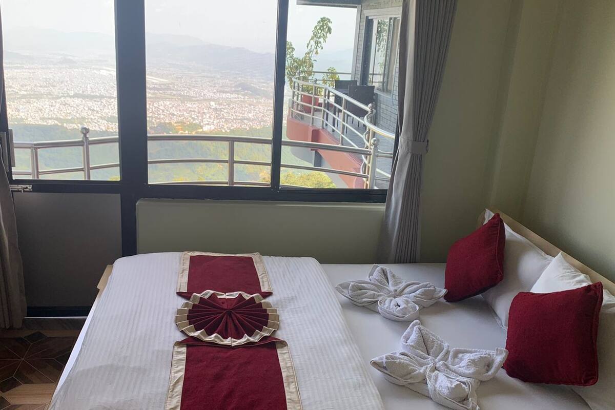 Habitación de hotel con un ventanal que ofrece vistas a la ciudad, una cama bien hecha con ropa de cama roja y blanca, y un balcón con barandilla.