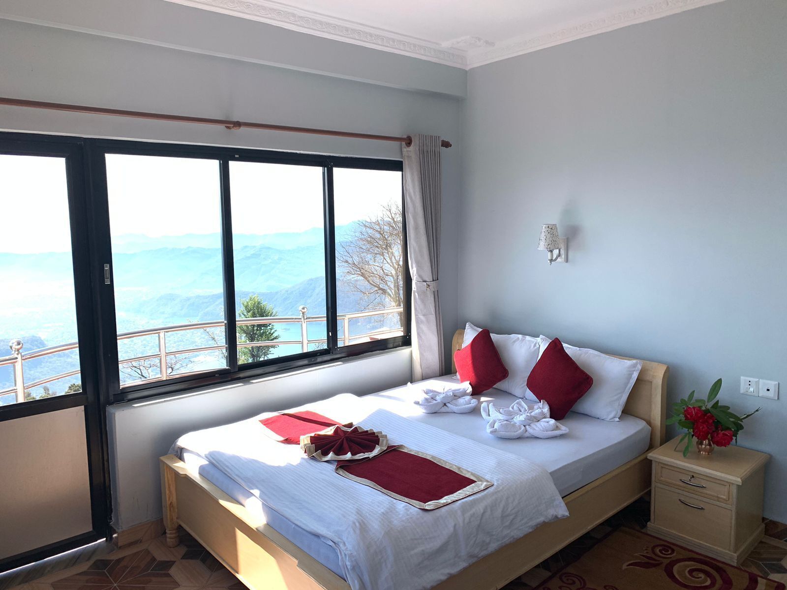 A hotel room with a large window offering a mountain view, a neatly made bed with red pillows, and a small bedside table with a vase of flowers.