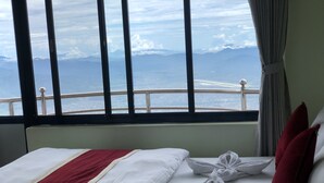 Ein Hotelzimmer mit Bett, Blick auf Meer und Berge durch große Fenster und einem Vorhang.