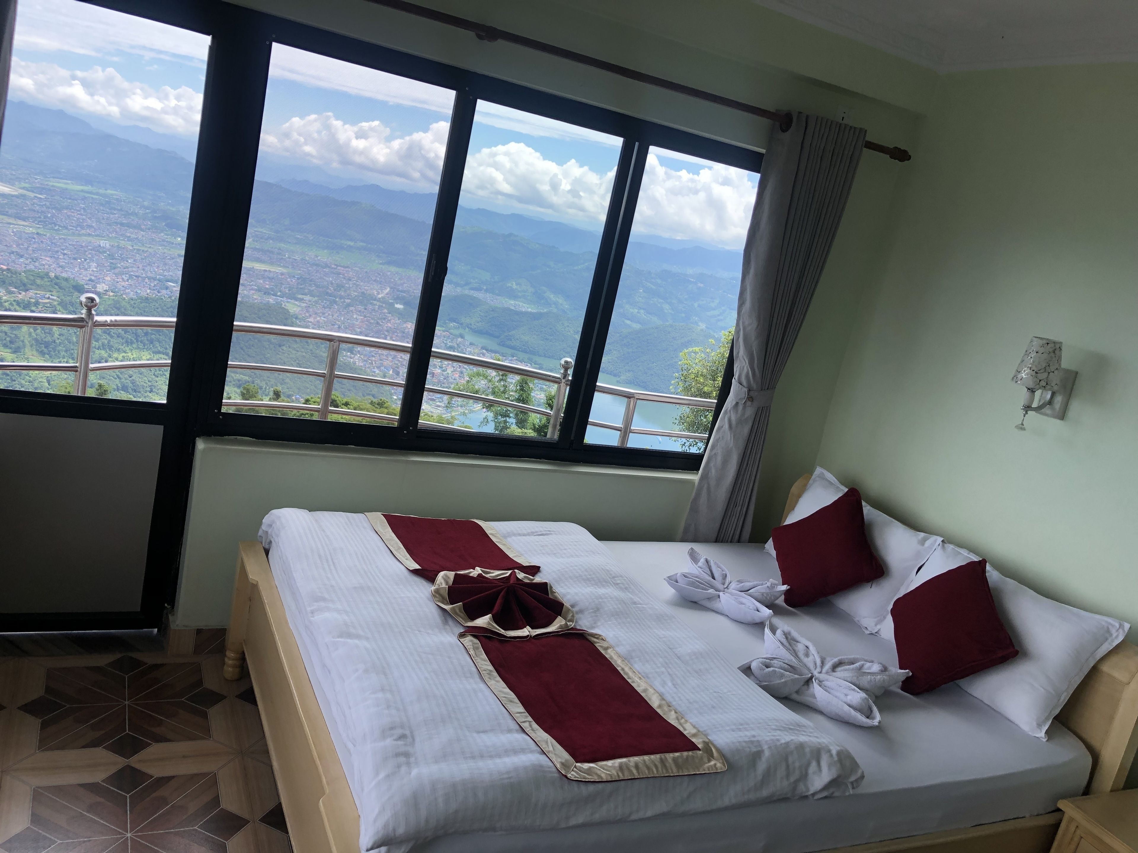 Une chambre d’hôtel dotée d’une grande fenêtre offrant une vue panoramique sur les montagnes et la ville. Le lit est parfaitement fait, avec du linge de maison blanc et des touches de couleur rouge.