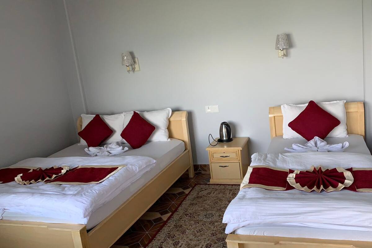 Habitación doble con dos camas, cada una con sábanas blancas y almohadas rojas, una mesita de noche de madera con hervidor, y una alfombra estampada.