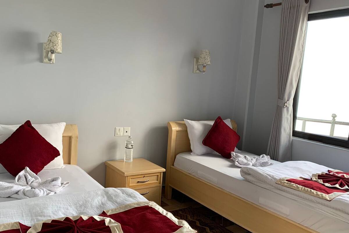 Habitación de hotel con dos camas individuales, cada una con cabecero de madera, ropa de cama blanca y almohadas rojas. Entre las camas hay una mesita de noche de madera con una botella de agua encima. La habitación tiene una ventana con cortinas y un aplique de pared.