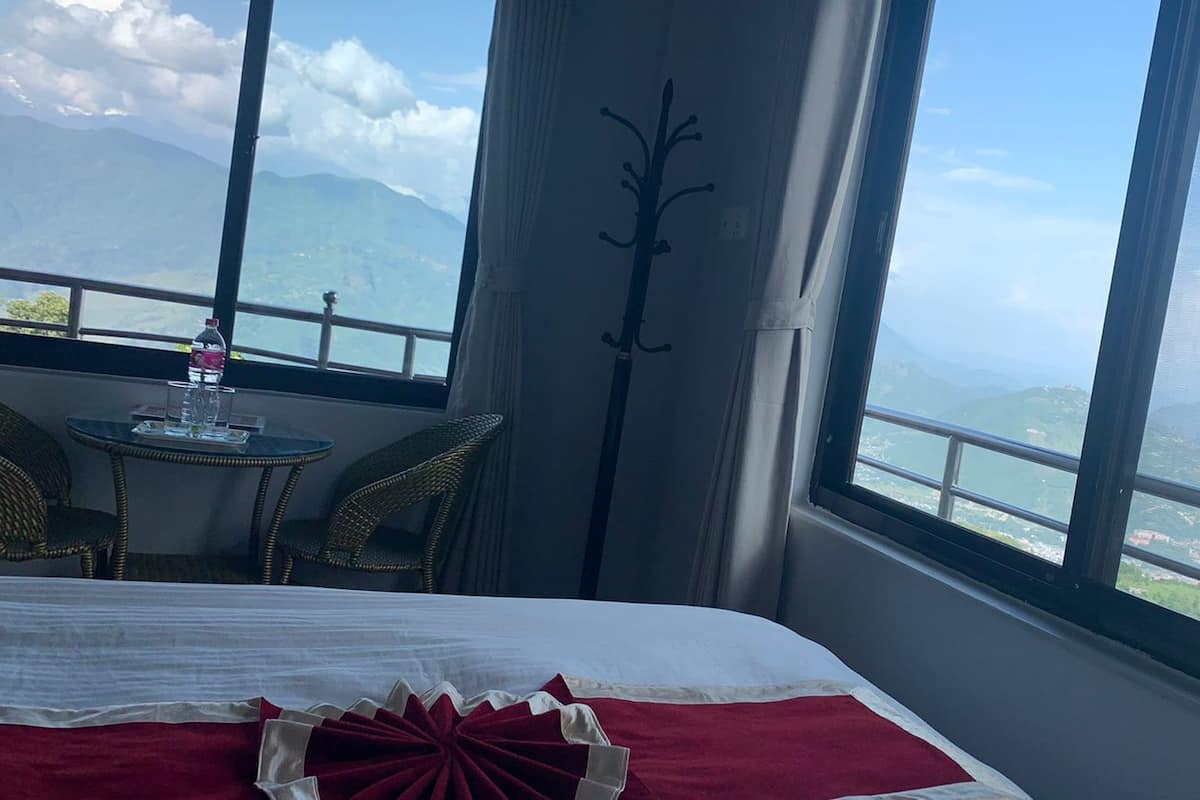 Habitación de hotel con cama, una mesa y sillas, y vistas a montañas a través de ventanas amplias.