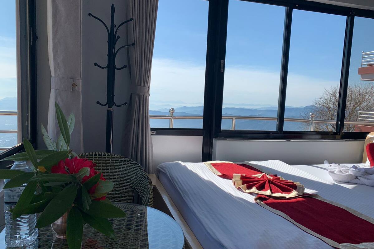 Habitación de hotel con una cama, una mesa con cubierta de vidrio y vistas a las montañas a través de amplios ventanales.