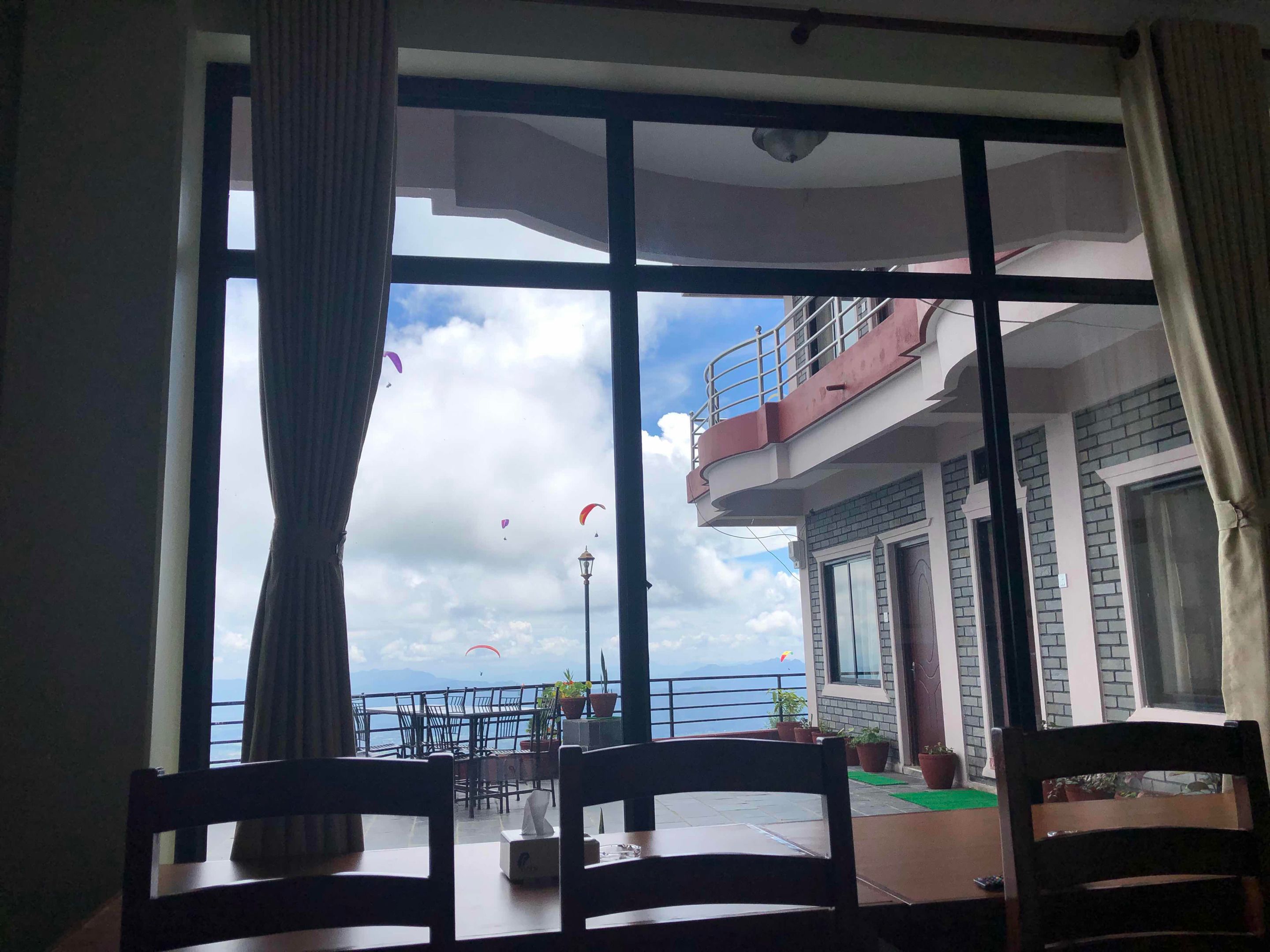 A room with a large window overlooking a balcony with potted plants and a view of the sea.