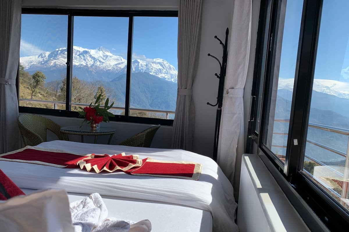 Habitación de hotel con una cama, una mesa con un jarrón de flores y vistas a montañas nevadas a través de amplias ventanas.