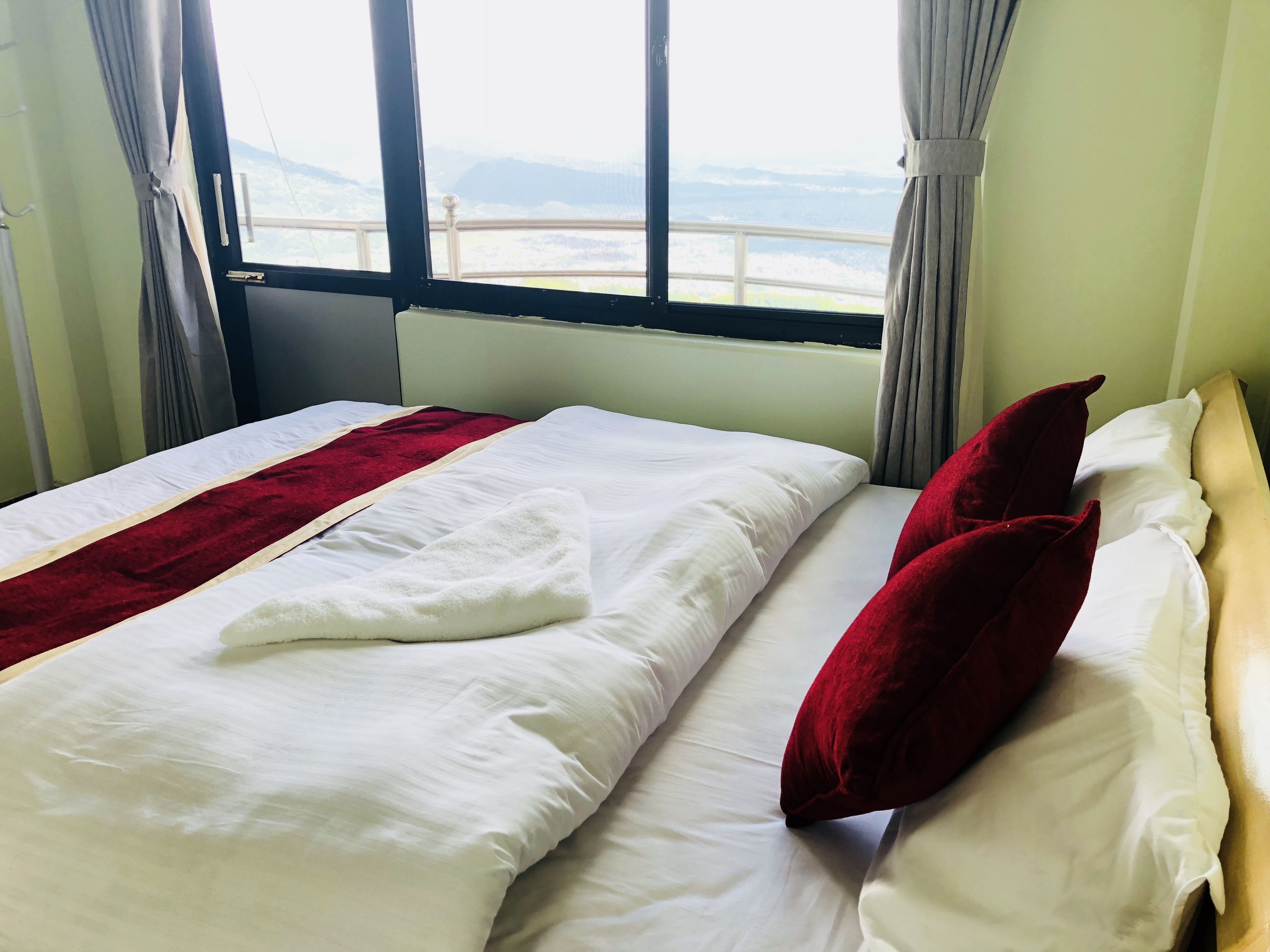 A neatly made bed with white and red bedding, a view of a distant landscape through a window, and a curtain on the left side.