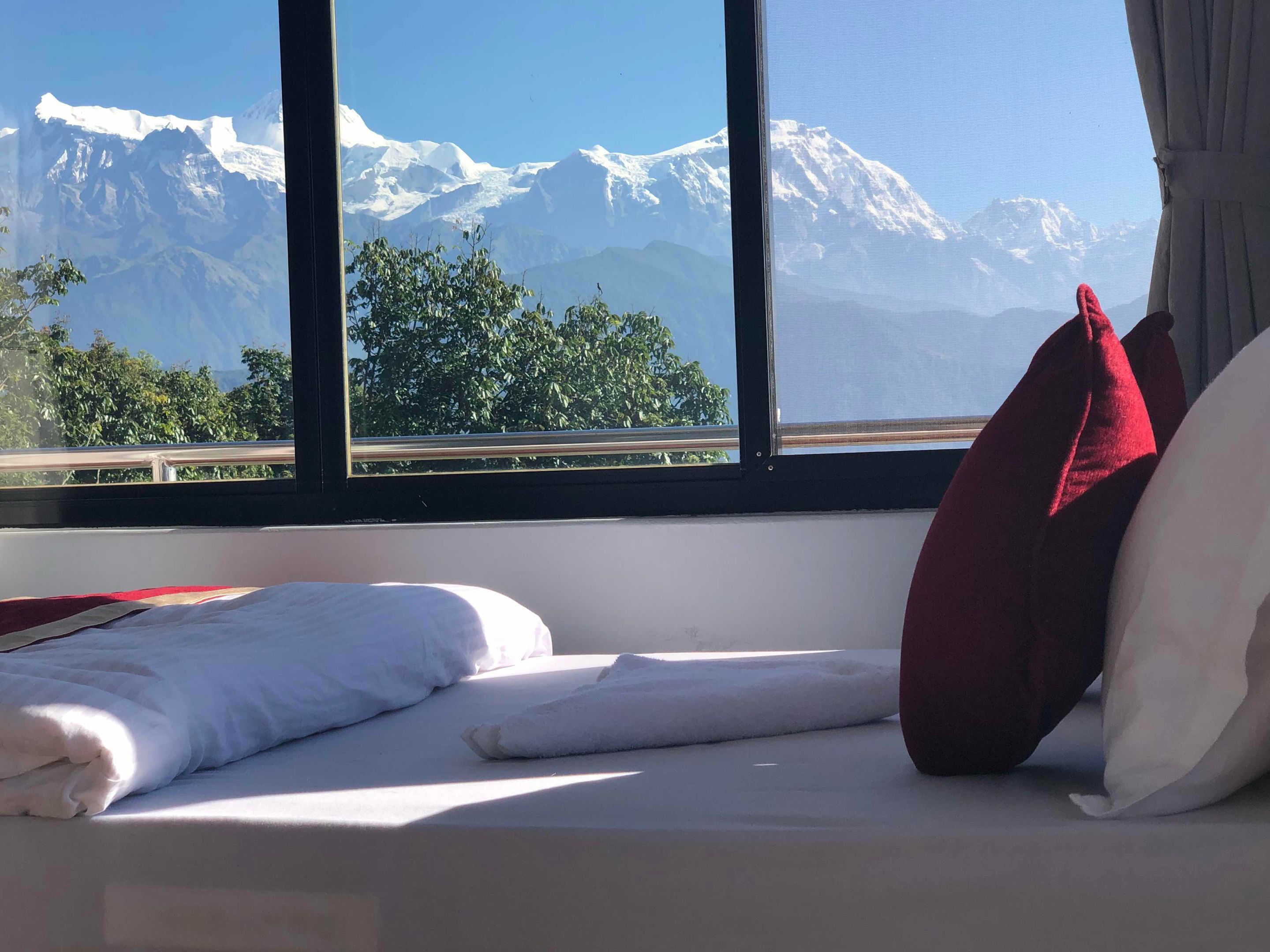 Une chambre d’hôtel avec un lit, du linge de lit blanc, un oreiller rouge et une vue sur des montagnes enneigées, visible à travers de grandes fenêtres.