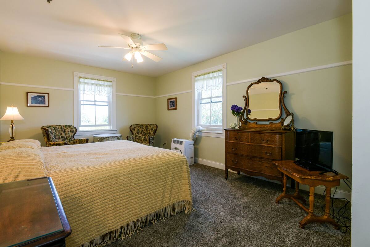 A bedroom with a bed, two chairs, a dresser, a television, and a ceiling fan.