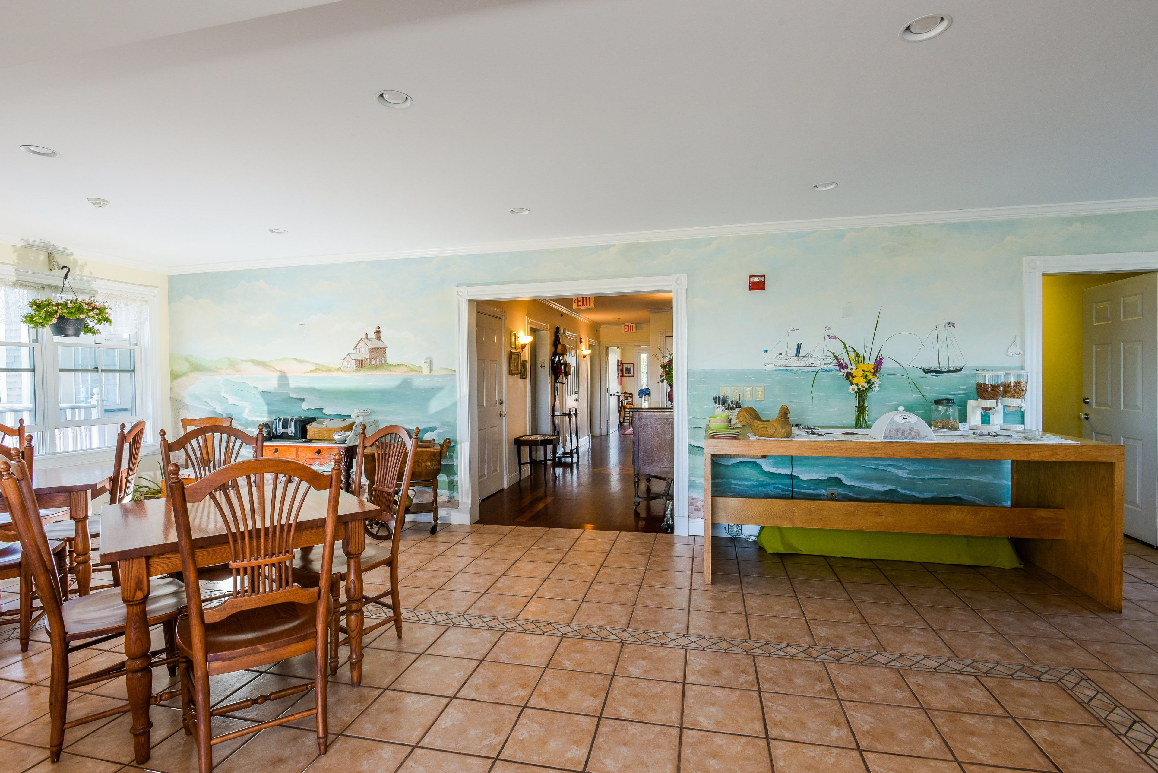 A dining area with wooden chairs and a table, a kitchen with a green countertop, and a mural of a coastal scene on the wall.