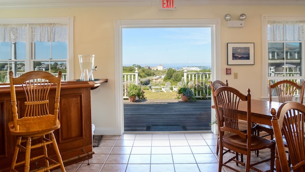 A dining area with a view of the landscape, wooden chairs and a table, and an exit sign above the door. - Payne's Harbor View Inn (Block Island)