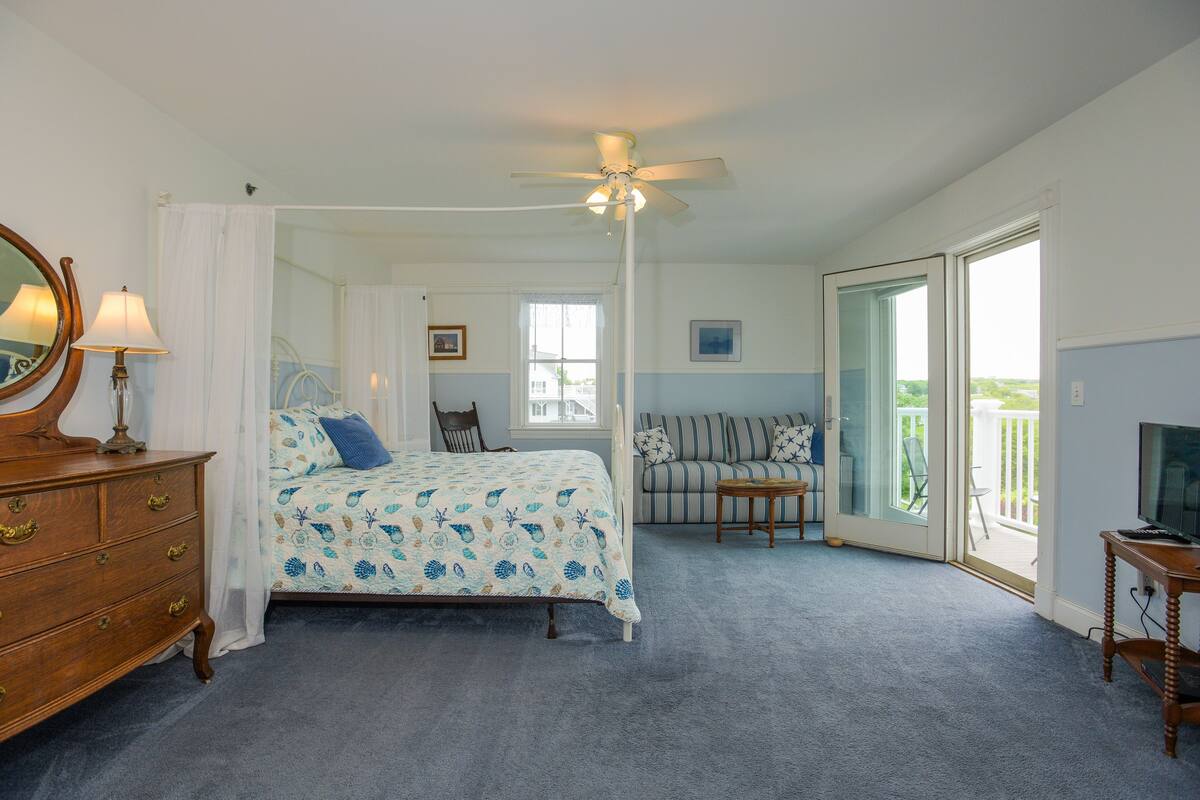 A bedroom with a bed, a dresser, a ceiling fan, a window, a sofa, and a television.