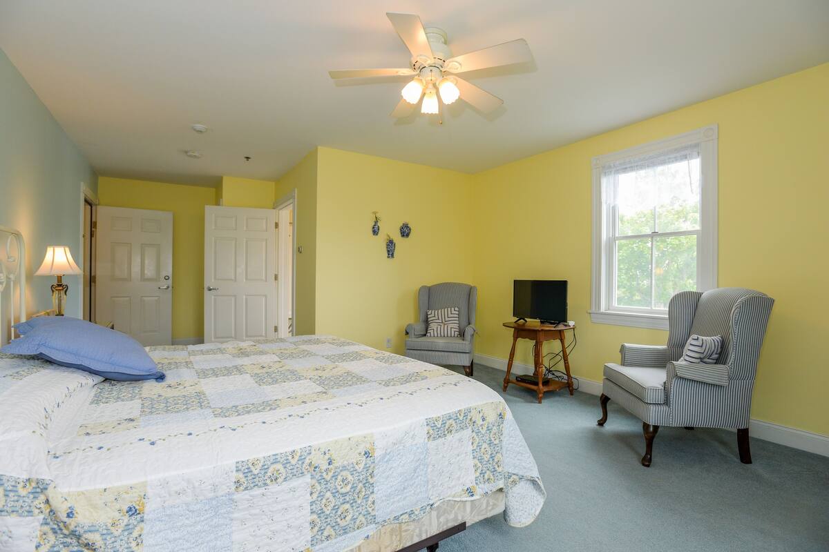 A bedroom with a bed, two chairs, a ceiling fan, a window, and a door.