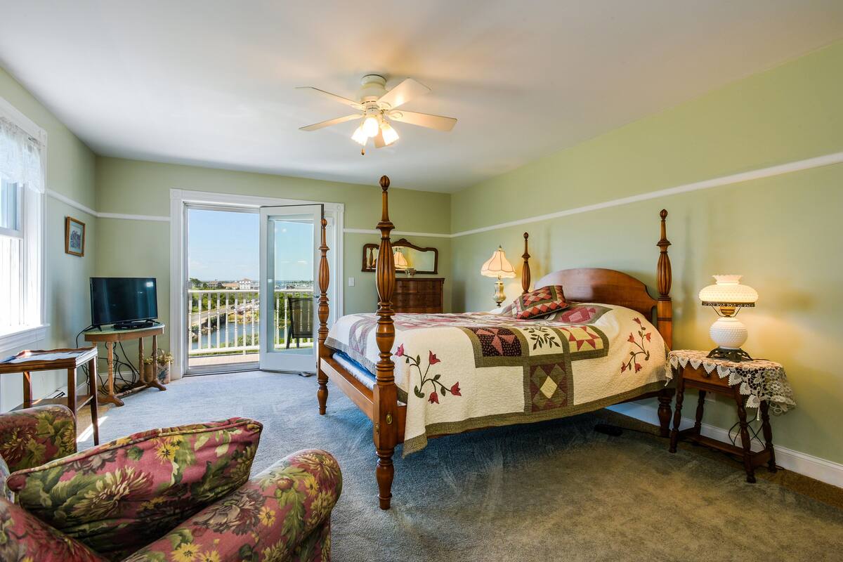 A bedroom with a four-poster bed, a television, a chair, and a balcony with a view.