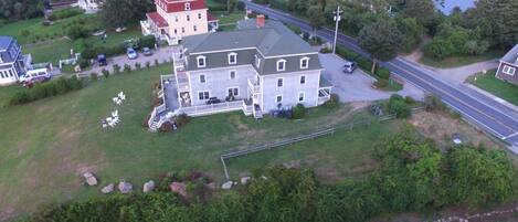 An aerial view of a large, multi-story house with a prominent red roof, surrounded by a well-maintained lawn and a road.