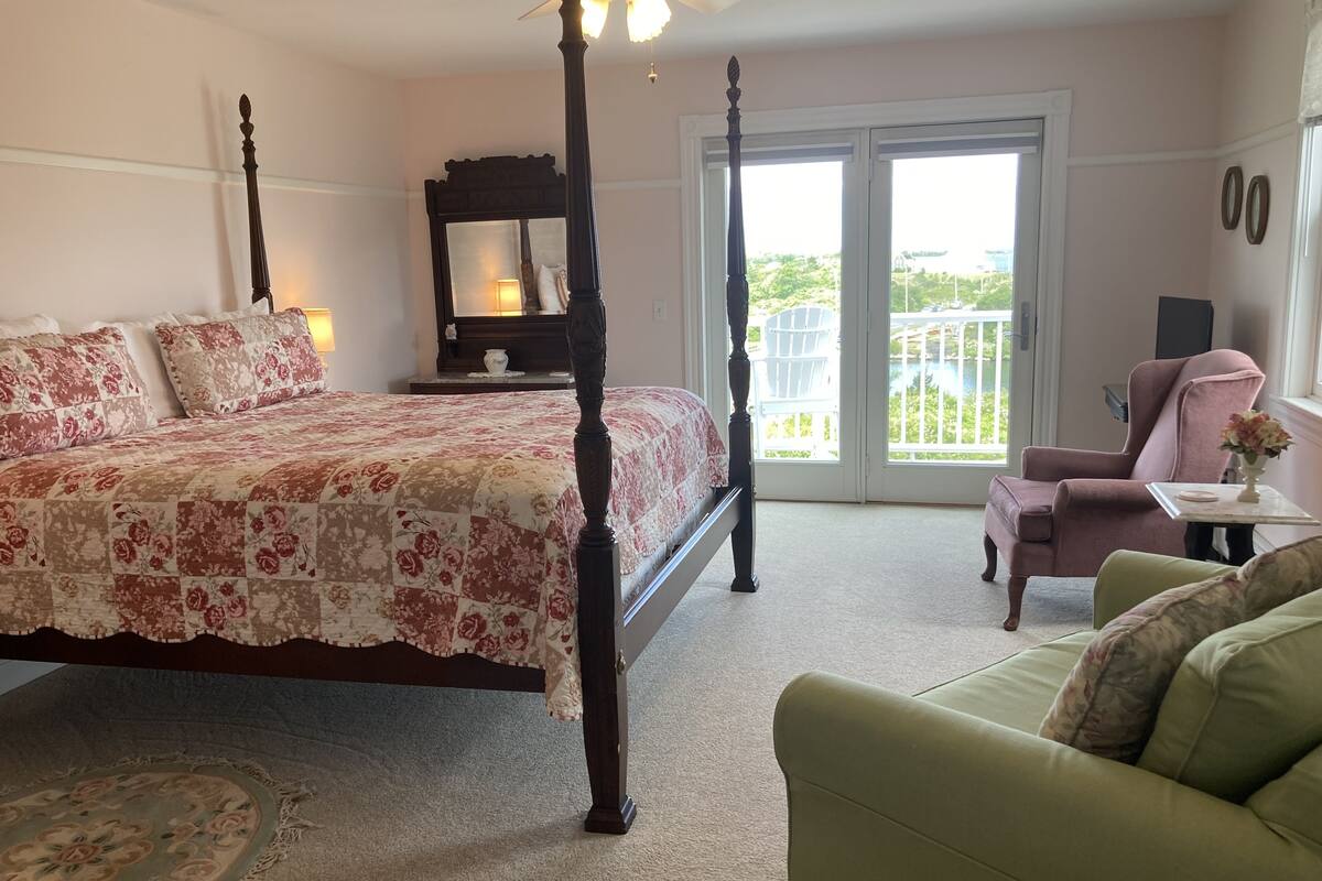 A bedroom with a four-poster bed, a green sofa, a small table with a vase, and a balcony with a chair.