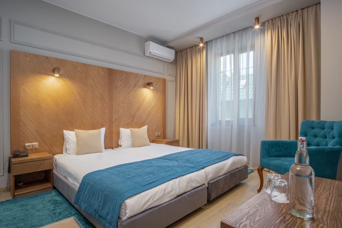 Een hotelkamer met een groot bed, een houten hoofdeinde, een blauwe accentmuur, een blauwgroene fauteuil, een houten bijzettafeltje en een fles water op het tafeltje.