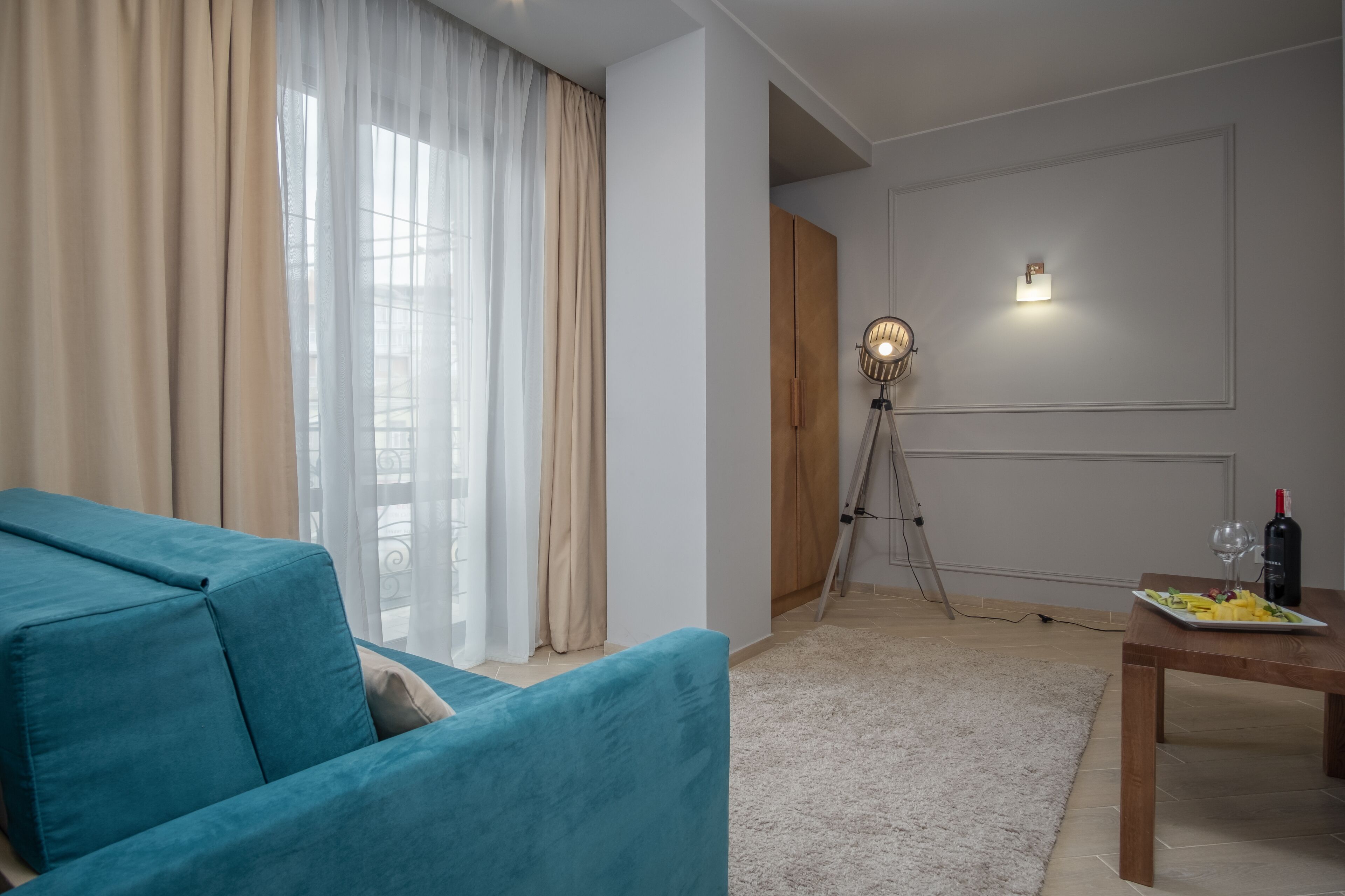 Ein Wohnzimmer mit einem petrolfarbenen Sofa, einem Esstisch mit einer Fruchtschale und einer Weinflasche, einer Dreibeinlampe und Blick durch transparente Vorhänge.