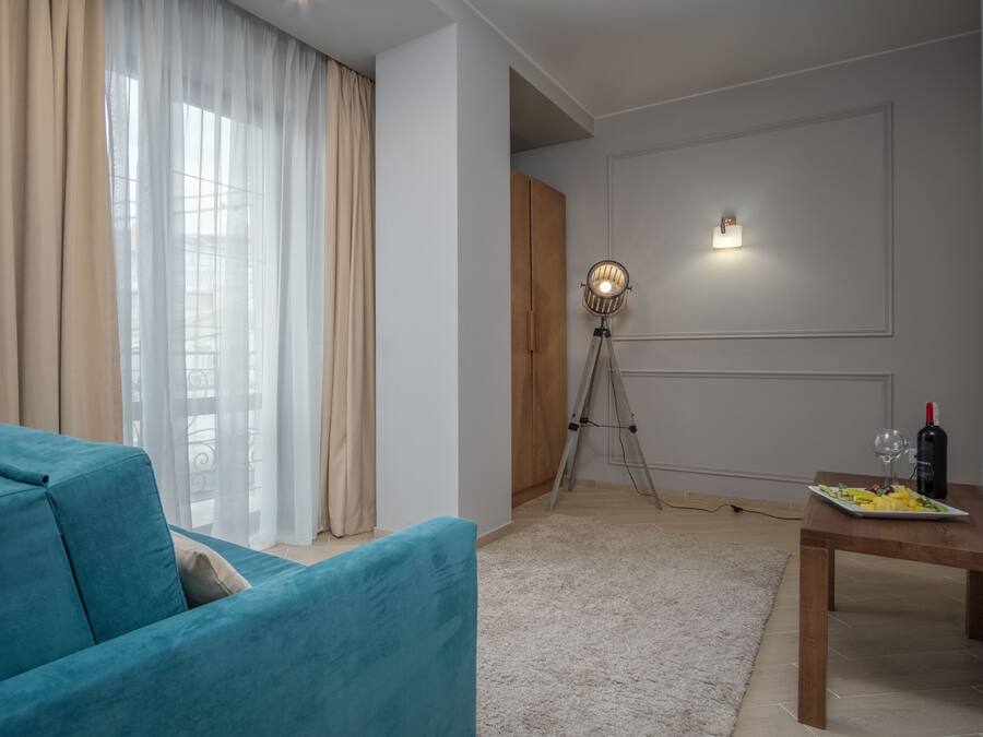 Ein Wohnzimmer mit einem petrolfarbenen Sofa, einem Esstisch mit einer Fruchtschale und einer Weinflasche, einer Dreibeinlampe und Blick durch transparente Vorhänge.