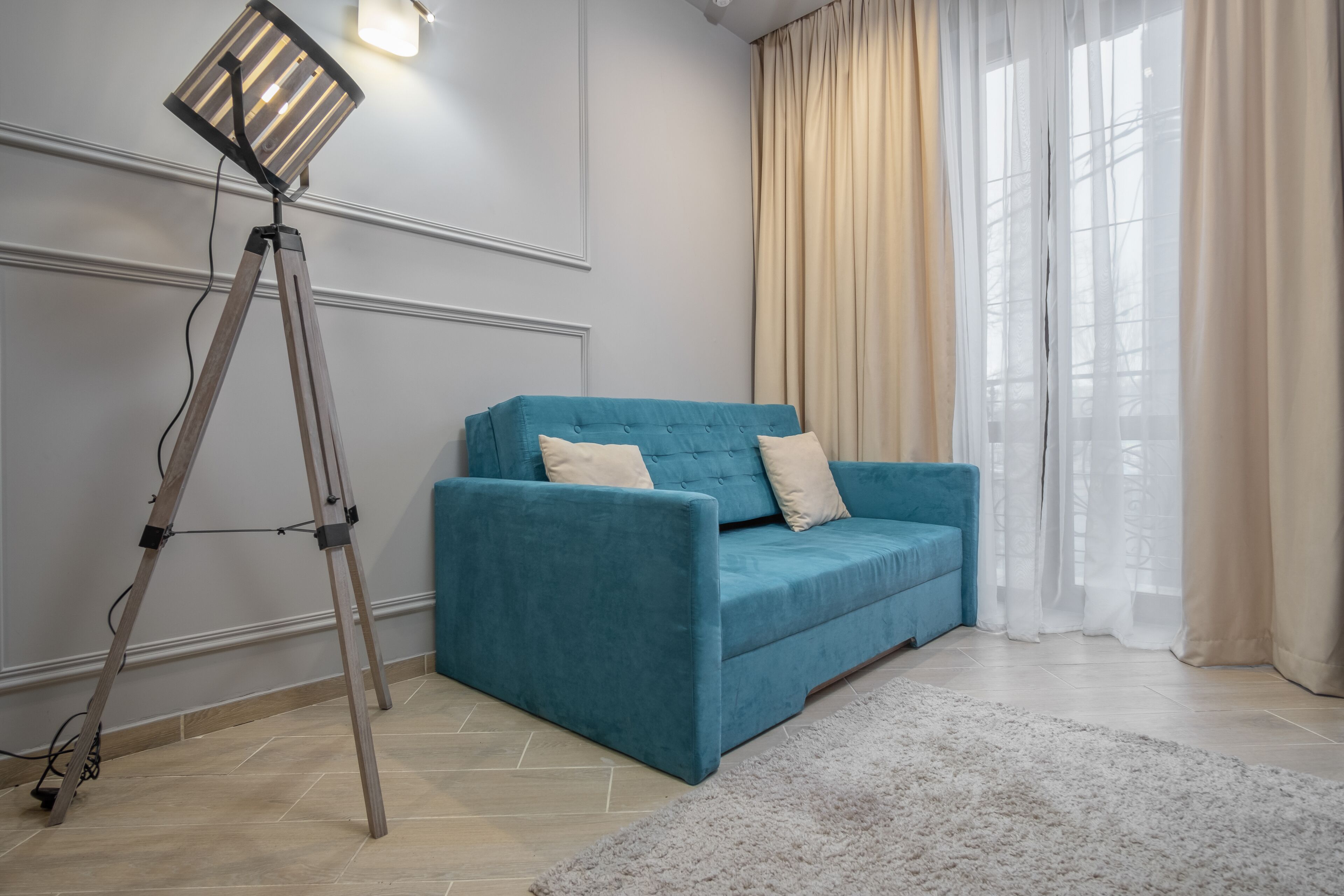 A modern living room with a teal sofa, a standing floor lamp, and a large window with sheer curtains.