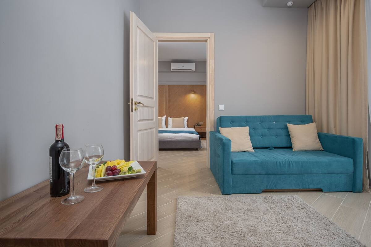 Een kamer met een houten tafel, een fles wijn en glazen, een blauwe bank en een bed op de achtergrond.