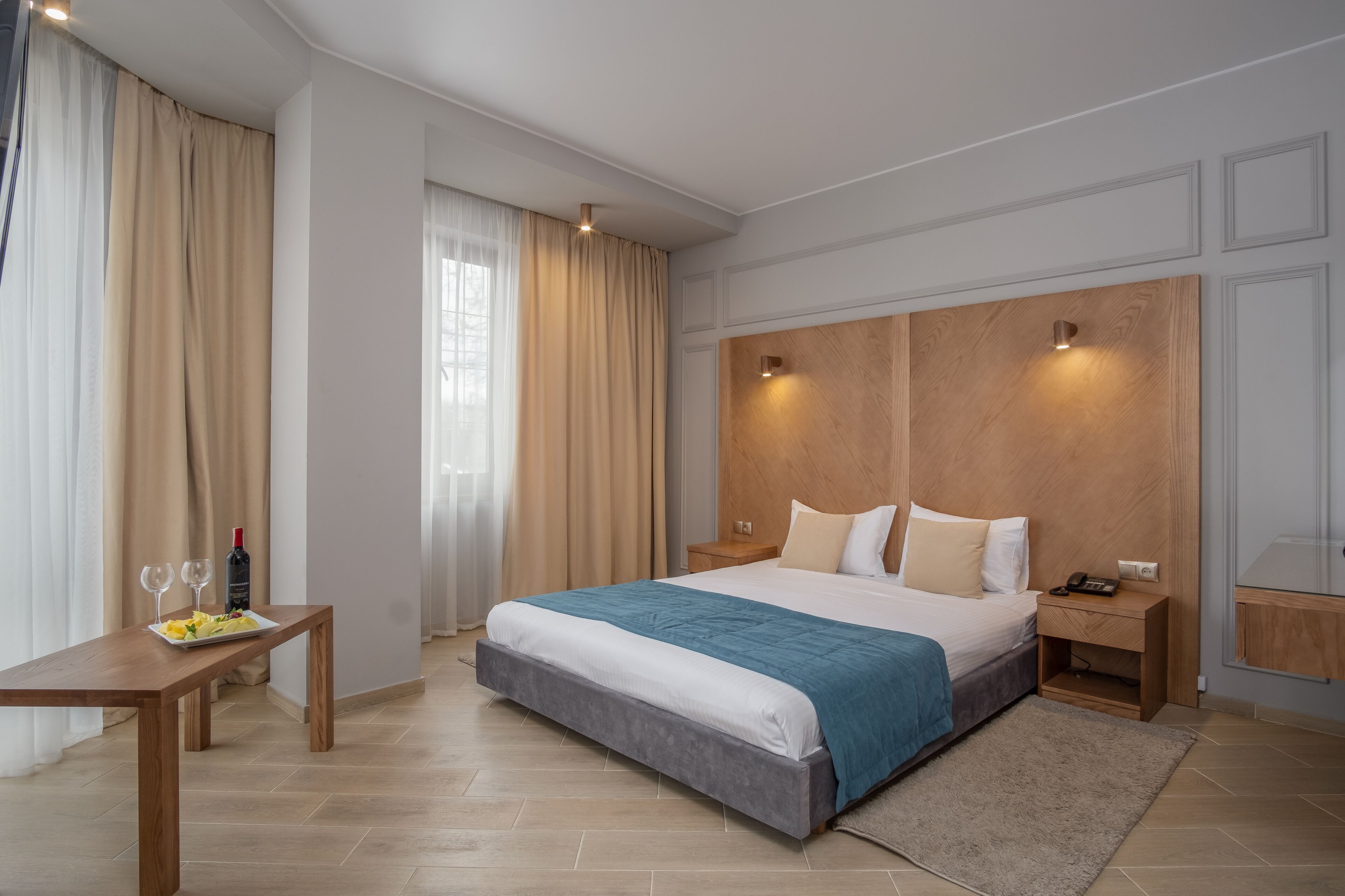 Une chambre d’hôtel moderne avec un grand lit, une tête de lit en bois, des tables de chevet et un petit bureau agrémenté d’une assiette de fruits et de verres à vin.