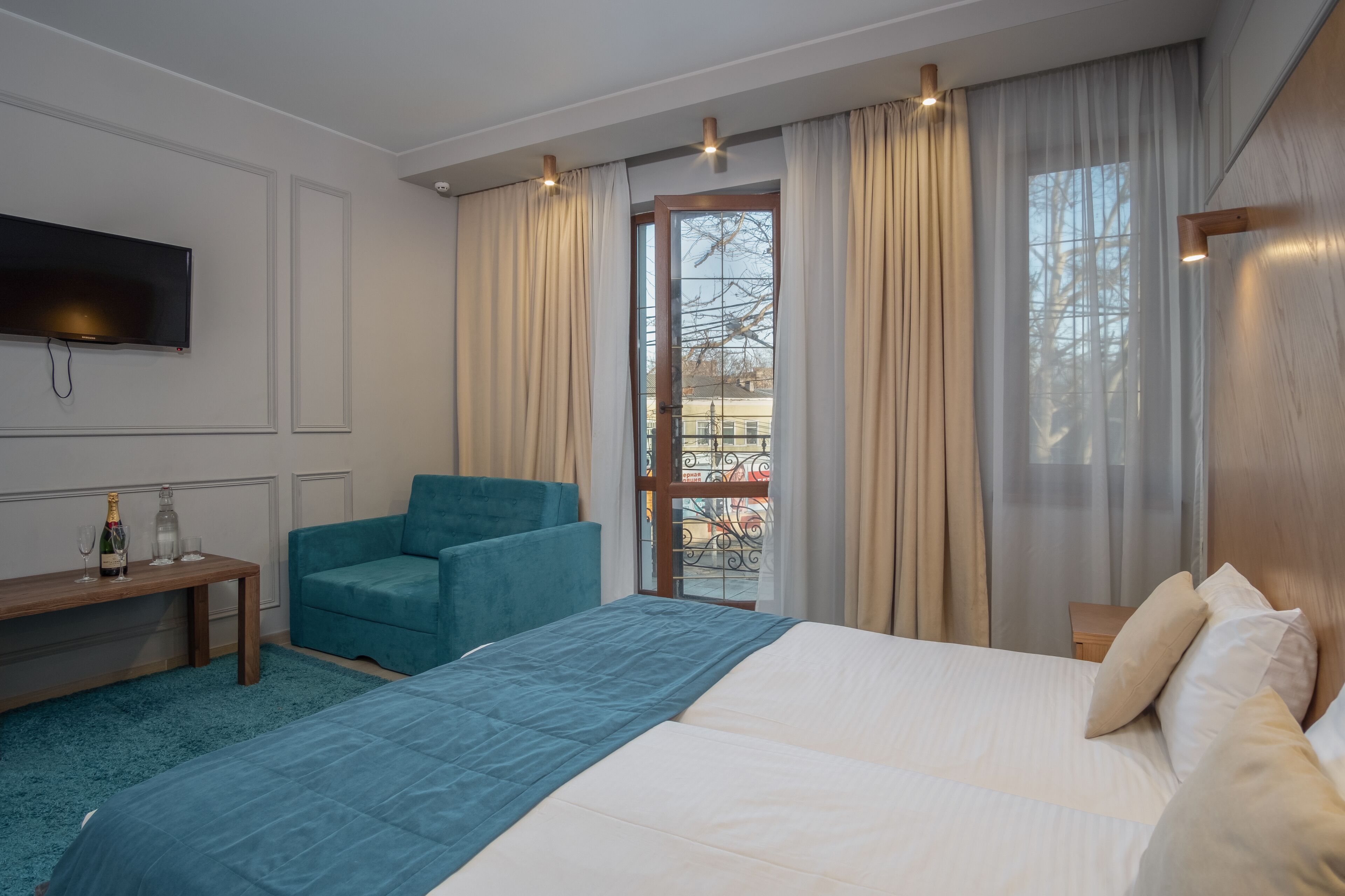 A hotel room with a large bed, a blue armchair, a wooden nightstand, a TV mounted on the wall, and a balcony with curtains.